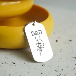 Sudraba krāsas metāla atslēgu piekariņš ar iegravētu uzrakstu DAD un ilustrāciju, kurā tēvs un bērns sadevušies rokās.