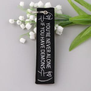 Mustast materjalist mootorratta võtmehoidja, mille servale on tikitud "You're never alone if you have demons".