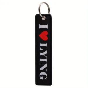 Medžiaginis pakabukas I LOVE LYING – juoda medžiaga su baltu išsiuvėtu užrašu ir raudonu širdies simboliu.
