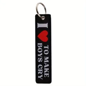 Medžiaginis pakabukas LOVE TO MAKE BOYS CRY – juoda medžiaga su baltu užrašu ir raudonu širdies simboliu.