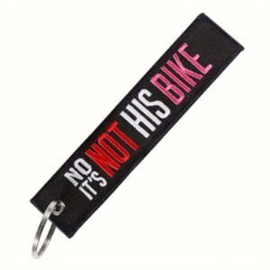 Must võtmehoidja, millele on tikitud valge, punase ja roosa tikandiga tekst "NO IT'S NOT HIS BIKE".