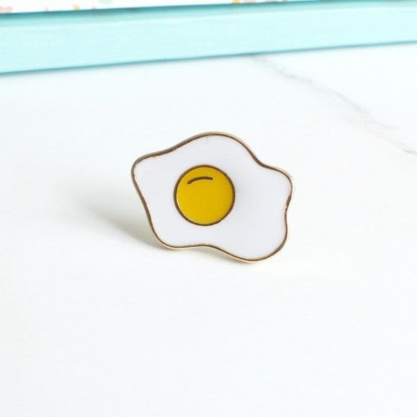White enamel egg cup with bright yellow yolk and gold outline pin accessory with a food theme