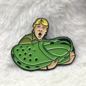 A blond-haired guy with a surprised expression holds a giant green Croc shoe enamel pin meme internet culture
