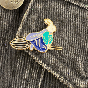 A white rabbit with a blue cloak flying on a witch's broomstick enamel pin fantasy fairy tales