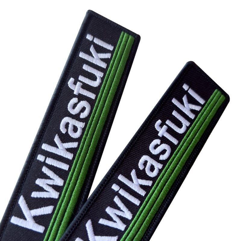 Du juodos spalvos tekstiliniai raktų pakabukai su baltu siuvinėtu užrašu „Kwikasfuki“ ir žaliomis detalėmis